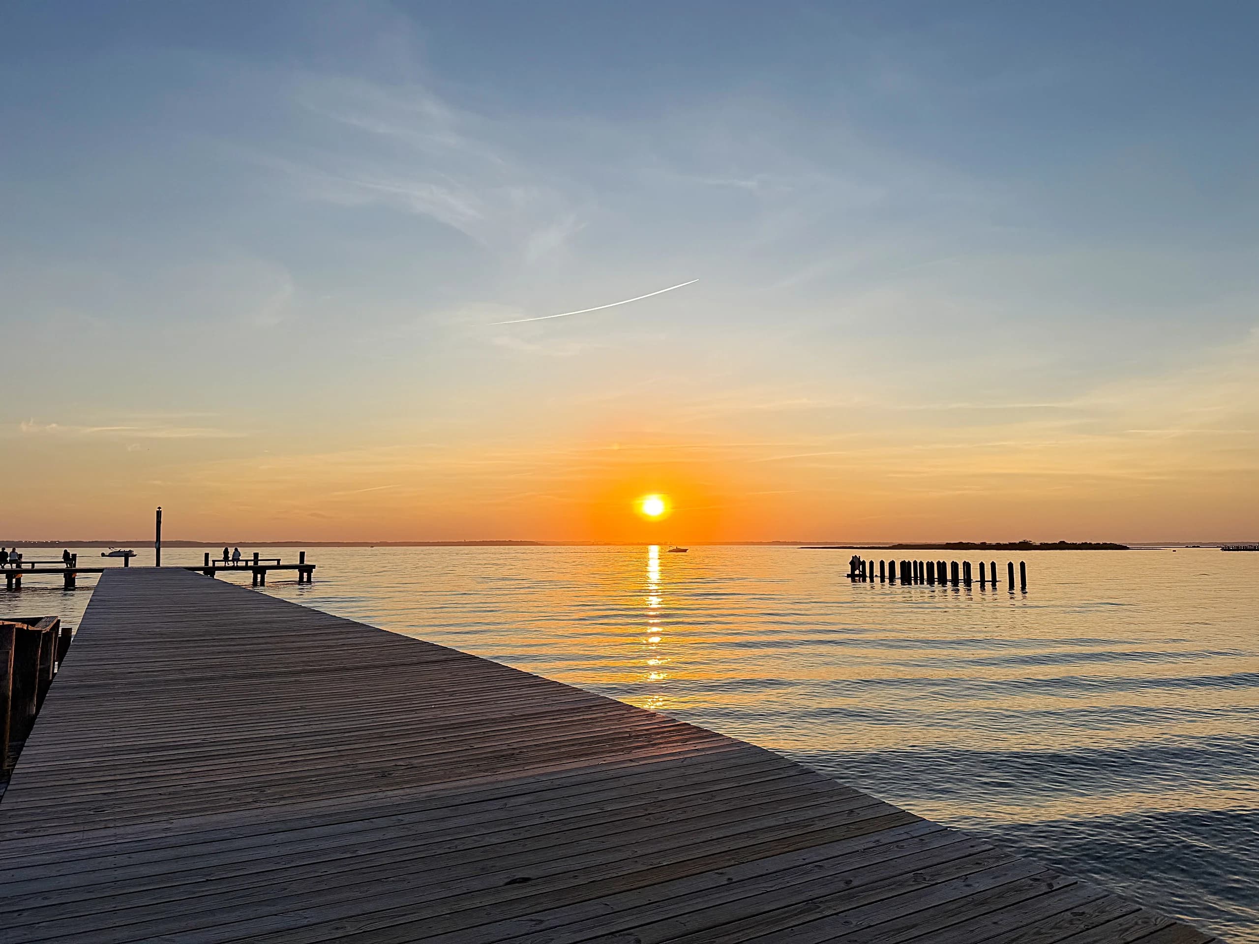 The sun setting over Barnegat Bay from the end of a long wooden dock, a boat crossing the light.